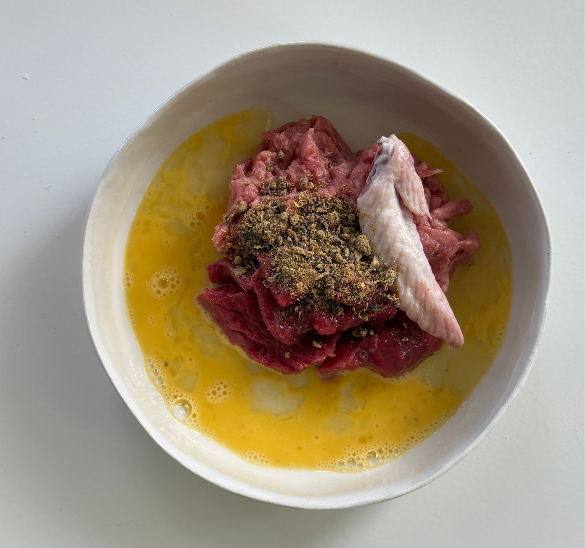A top-down view of raw meat, including a chicken wing tip, mixed with beaten eggs and Essential Blend for cats
 in a white bowl.