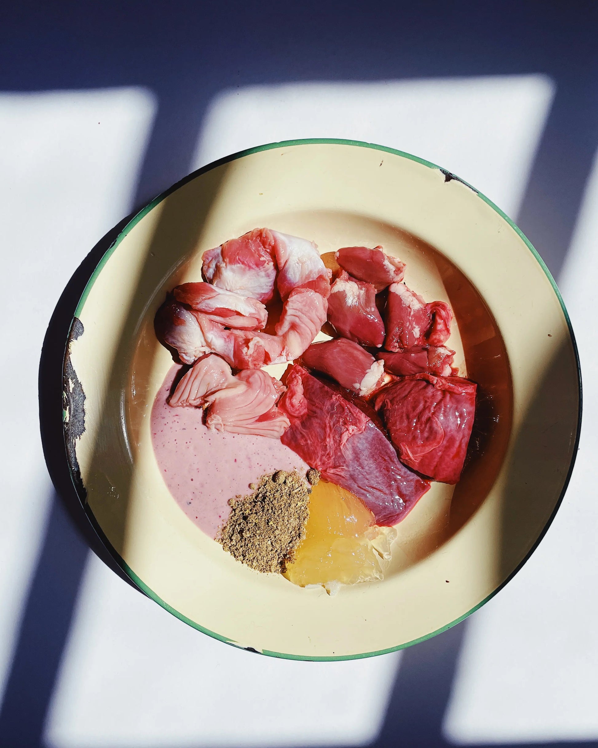 Raw meat pieces, a yellow gelatin cube, and a spoonful of brown Essential Blend for dogs powder, arranged in a round cream-colored enamel bowl with a green rim, set against a background with strong window shadows.