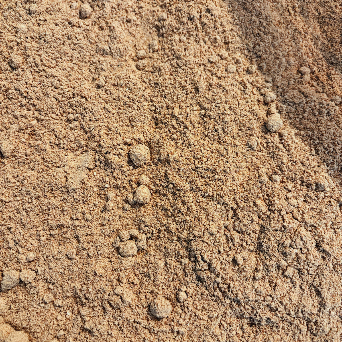A close-up view of a textured pile of fine brownish-tan powder or granules with small lumps, filling the entire frame.