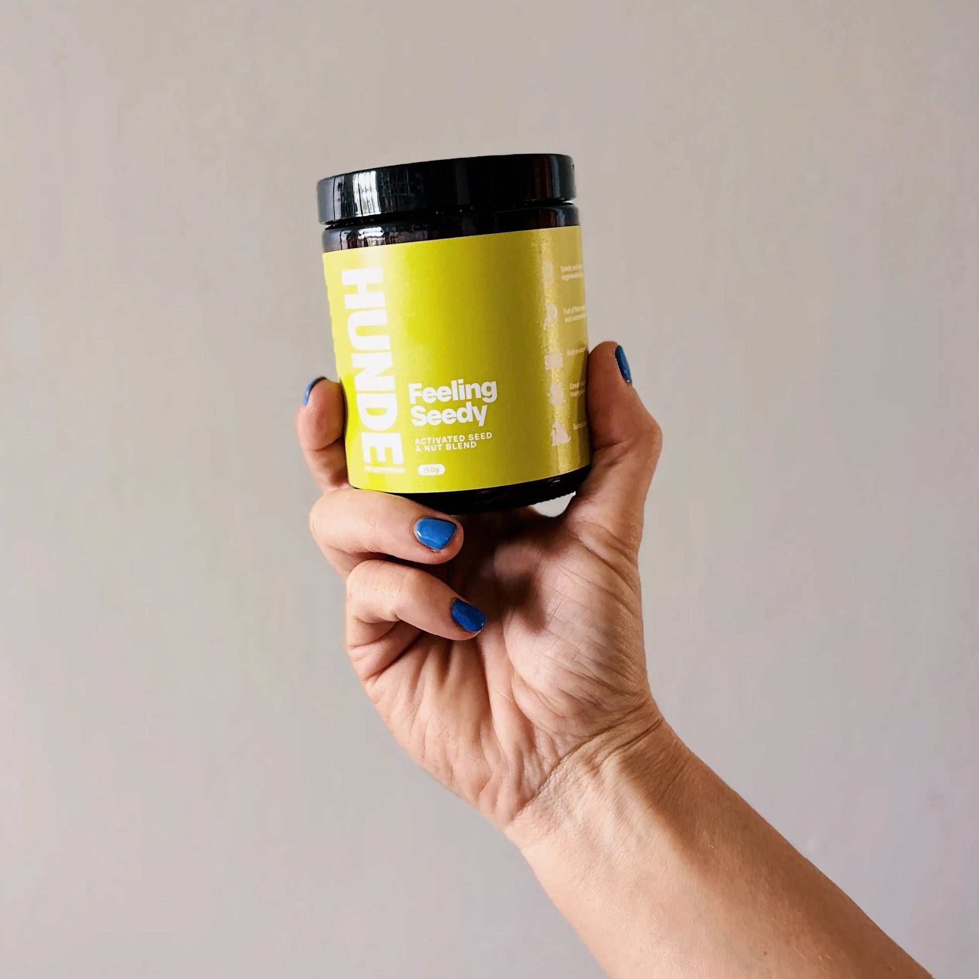A hand with blue nail polish holds an amber jar of Hunde Feeling Seedy supplement against a plain beige wall.