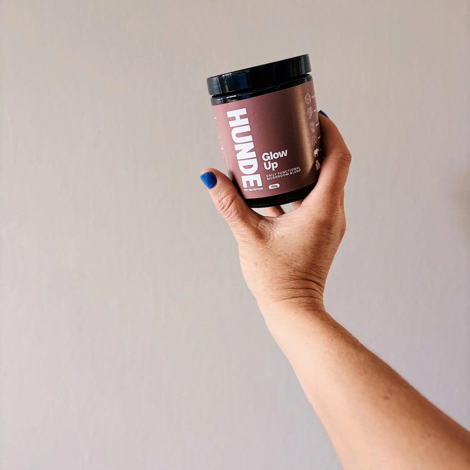 A hand with blue nail polish holds an amber jar of Hunde Glow Up powder against a plain wall.
